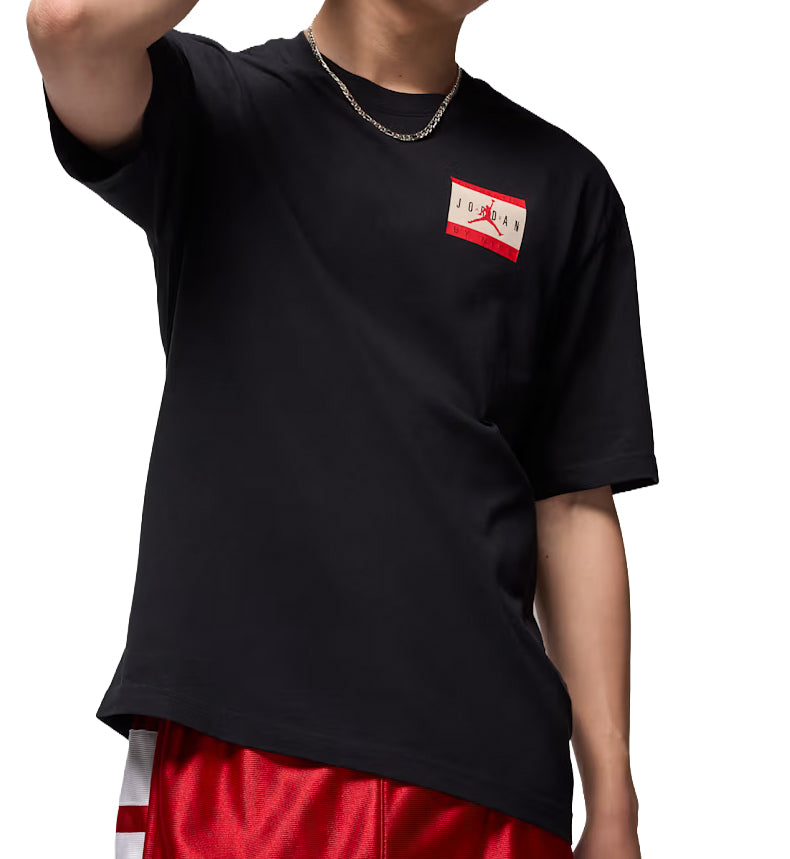 Person wearing a black Jordan Sportswear T-Shirt with a red and white logo on a plain background